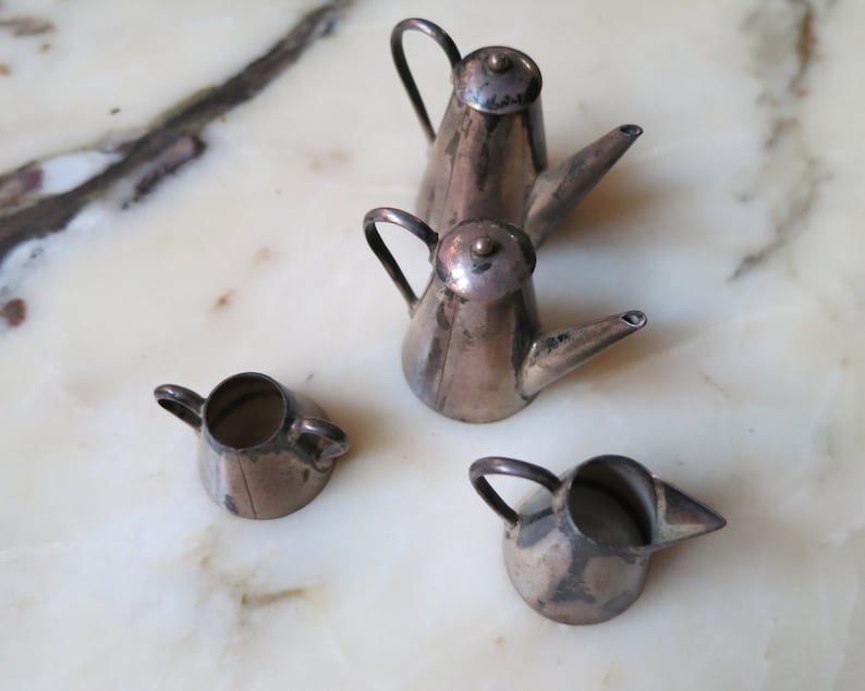 May include: Four silver miniature teapots with handles and spouts. The teapots are arranged on a white marble surface.