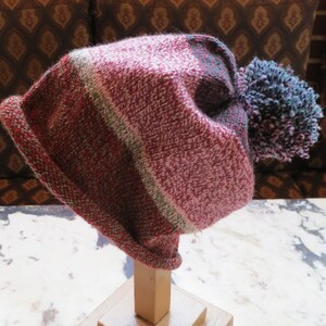 May include: A pink, green, and gray striped knit beanie with a fluffy blue and purple pom pom.