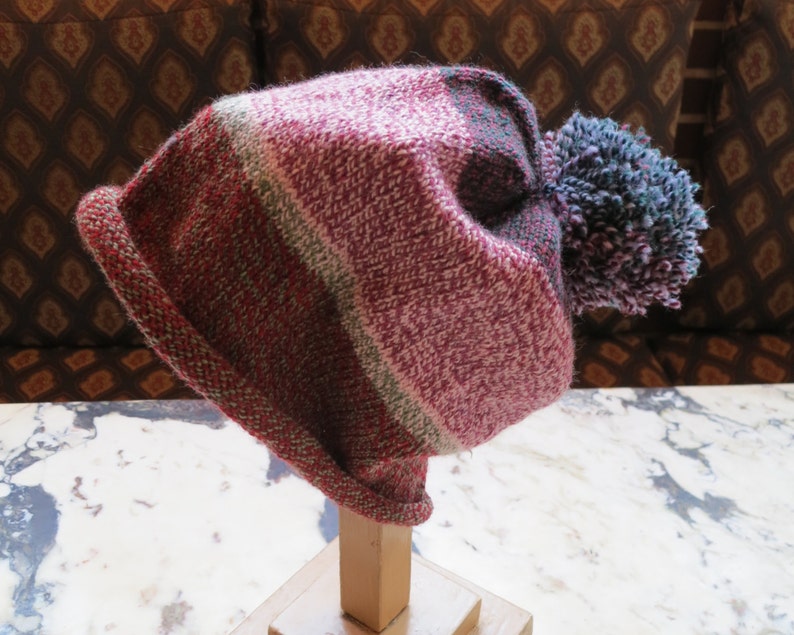 May include: A pink, green, and gray striped knit beanie with a fluffy blue and purple pom pom.