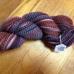 May include: A skein of yarn in a multi-colored tweed pattern with shades of red, pink, purple, green, and black. The yarn is twisted into a rope shape and has a white tag attached with a barcode.