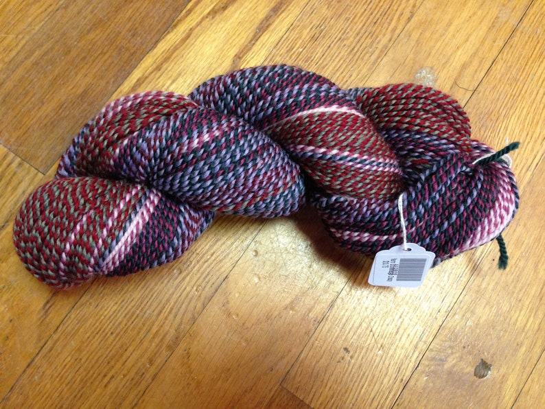 May include: A skein of yarn in a multi-colored tweed pattern with shades of red, pink, purple, green, and black. The yarn is twisted into a rope shape and has a white tag attached with a barcode.