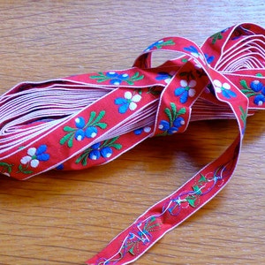 May include: Red ribbon with white and blue floral embroidery. The ribbon is folded and tied in a knot. The ribbon is on a wooden surface.