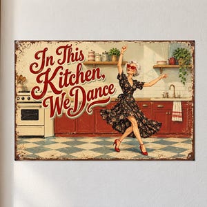 May include: A rectangular metal sign featuring a kitchen scene with a person dancing in a floral dress. The text "In This Kitchen, We Dance" is written in large red script. The background shows red cabinets, a stove, and a checkered floor.