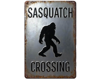 May include: A rectangular metal sign with the words "SASQUATCH CROSSING" in black, with a black silhouette of Bigfoot walking across the center. The sign has a distressed, rusty border.