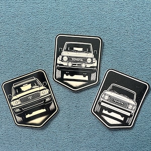 May include: Three black and white Toyota SUV stickers on a blue textured surface. Each sticker features a different front view of a Toyota SUV model. The stickers are shield-shaped with a black border and white background.
