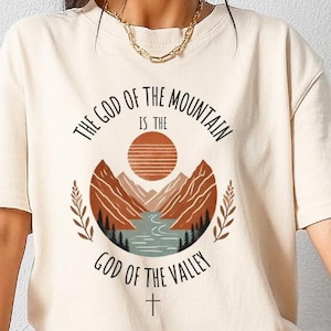 May include: Cream-colored t-shirt featuring a graphic design with the text "THE GOD OF THE MOUNTAIN IS THE GOD OF THE VALLEY" in a circular arrangement. The design includes a mountain range, sun, river, and cross.
