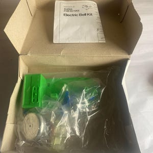 May include: An open cardboard box containing an Electric Bell Kit. The kit includes a green plastic component, a small wooden wheel, and other clear plastic parts, all sealed in a plastic bag. A white instruction sheet is visible.