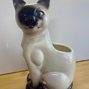 May include: A ceramic Siamese cat figurine planter with a glossy cream-coloured body and dark brown points on the ears, face, paws, and tail. The cat is in a seated position with an open cavity on its back for holding small items or plants.