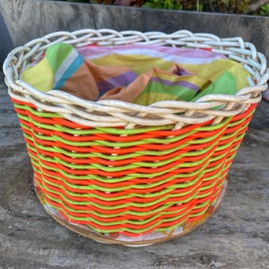 May include: A round woven basket with a white rim and a colourful orange and green body. The basket contains a colourful striped fabric. The basket is sitting on a wooden surface.
