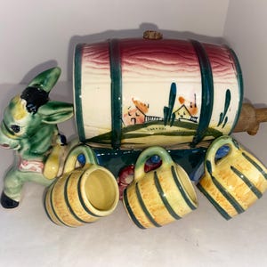 May include: A ceramic donkey pulling a barrel-shaped container with three small mugs. The barrel is red and white with a landscape design. The mugs and barrel have green and yellow stripes.