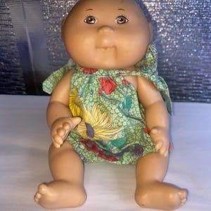 May include: A vintage baby doll with a bald head and brown eyes. The doll is wearing a green floral print dress with yellow and red flowers. The doll is sitting with its legs extended.