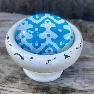 May include: A white, distressed cabinet knob with a blue and white patterned glass insert. The knob has a rounded shape and a pedestal base, suitable for furniture or decorative projects.