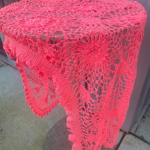 May include: A bright neon pink circular crochet tablecloth featuring an intricate floral pattern with repeating sunflower-like motifs and a delicate scalloped edge. The textile is draped over a round surface, showcasing the openwork lace design.