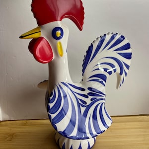 May include: A ceramic rooster figurine with a red comb and wattle, yellow beak, and blue and white patterned feathers. The base is white with blue accents. The rooster is a decorative item.