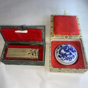 May include: Two decorative boxes with red velvet interiors. One contains a rectangular stone seal with carved details. The other holds a round, white ceramic seal with a blue dragon design.
