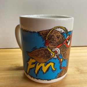 May include: White ceramic coffee mug featuring a cartoon bear wearing red goggles and a yellow diving mask. The mug has a blue background and yellow text that reads "FM".