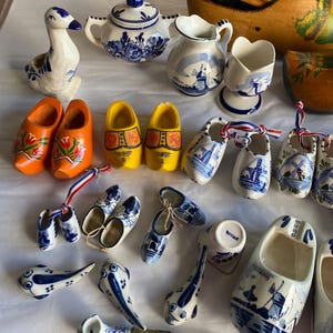 May include: A collection of ceramic and wooden Dutch-themed souvenirs including a duck figurine, a sugar bowl, a pitcher, a cup, and numerous miniature wooden clogs. Several items feature blue and white windmill patterns, while others are painted in solid orange or yellow with floral designs. A long, thin pipe with a blue and white ceramic bowl is also included.
