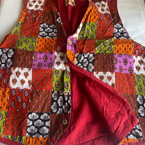 May include: A colourful patchwork vest with a variety of patterns. The front is made up of squares in orange, red, brown, green, purple, and white, each with floral and paisley designs. The vest has a red lining and wooden buttons.
