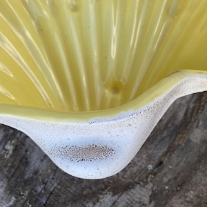 May include: A close-up of a yellow ceramic dish with a scalloped design. The dish has a white, textured rim. The interior of the dish has vertical ridges. The dish is likely vintage or antique.
