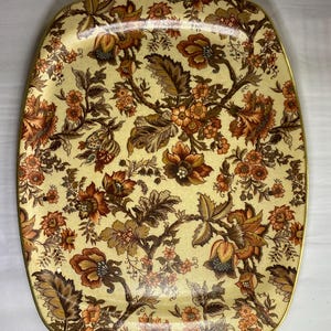 May include: A rectangular serving tray with rounded corners, featuring a cream-coloured background decorated with a dense, repeating floral pattern in shades of burnt orange, brown, and muted gold, finished with a thin gold-toned metal rim.