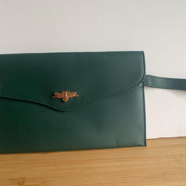 Könnte beinhalten: Eine dunkelgrüne Clutch mit Klappenverschluss und kleinem Griff. Die Clutch hat ein goldfarbenes Bienenemblem auf der Vorderseite. Die Clutch ist aus einem glatten Material gefertigt.