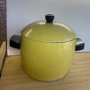 May include: A round, chartreuse yellow cooking pot with a matching lid and two black side handles. The pot features a smooth, solid-coloured exterior finish with a small black knob on the centre of the lid and a metallic base rim.