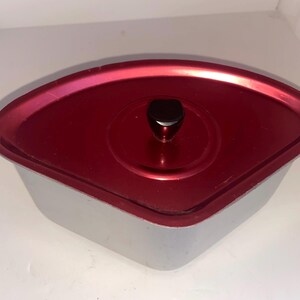 May include: A silver-coloured, lidded container with a red top. The lid has a black knob in the centre. The container has a unique, angular shape, and the lid appears to be made of a different material than the base.