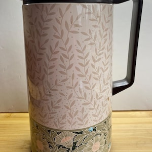 May include: A vintage insulated flask with a black handle, spout, and lid. The body has a cream background with a repeating pattern of tan leaves and dots. The bottom has a floral design with green, tan, and pink accents.