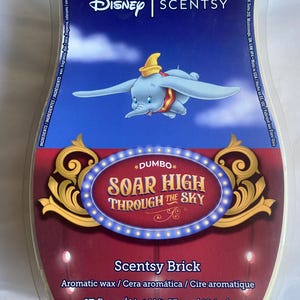 May include: A Scentsy Brick featuring Dumbo the elephant soaring through the sky. The wax brick has a blue and red background with gold accents. It contains 503 ml of aromatic wax, with a net weight of 424 g.