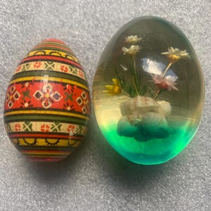 May include: Two decorative Easter eggs. One is wooden, painted with a red, yellow, and green geometric pattern. The other is clear, with flowers and a green and blue base. A unique Easter decoration.