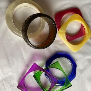 May include: A collection of seven bangle bracelets in various shapes and colours, including a clear round bangle, a dark brown textured round bangle, a red square bangle, a yellow square bangle, a blue hexagonal bangle, a green square bangle, and a purple square bangle.