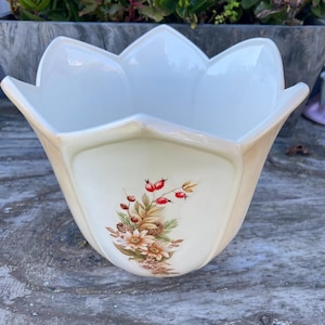 May include: Cream ceramic planter with a floral design. The planter has a tulip-like shape with a scalloped rim. The floral design features red berries, pinecones, and beige flowers. The planter is sitting on a weathered wooden surface.