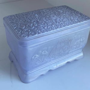 May include: A pale lavender trinket box with an ornate design and a ship scene on the front. The box has a scalloped edge and a raised lid.
