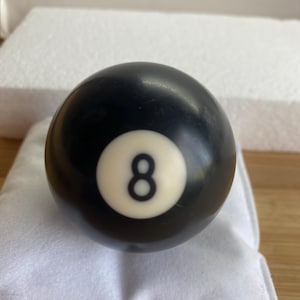 May include: A spherical black billiard ball featuring a centred white circular inlay with the black number eight printed in the middle. The surface of the ball is smooth and glossy, reflecting light across its dark, rounded exterior.