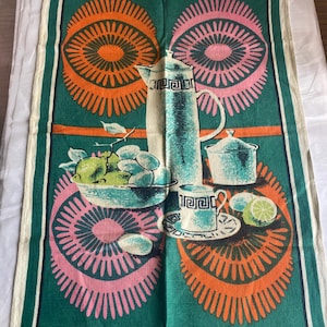 May include: A rectangular tea towel featuring a dark green background with large orange and pink sunburst patterns. The central graphic depicts a tall pitcher, a lidded sugar bowl, a teacup on a saucer, and a bowl filled with green citrus fruits.