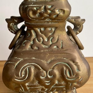 May include: A vintage bronze vase with detailed relief carvings and two ring handles. The vase has a bulbous body and a flared top. The bronze has a patina with hints of green.