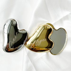 以下が含まれることがあります： 白いサテンの表面に、銀色と金色の2つのハート型の美容ツールが置かれています。銀色のツールには「Diara」、金色のツールには「Megan」と刻まれています。これらはスキンケア用に設計されています。