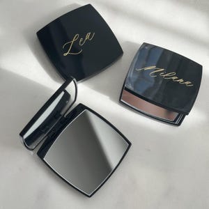 Op de afbeelding: Drie zwarte compacte spiegels met gouden scriptletters. Eén spiegel is open en onthult een reflecterend oppervlak. De andere twee spiegels zijn gesloten, met de namen "Lea" en "Milana" in gouden script. De spiegels hebben een vierkante vorm met afgeronde hoeken.