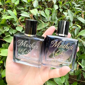 Op de afbeelding: Twee vierkante glazen parfumflesjes met zwarte doppen. De flesjes hebben een kleurenschema met verloop, dat overgaat van donkerblauw naar lichtroze. De woorden "Mother of the Bride" staan in elegant schrift op de voorkant.