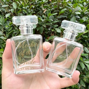 May include: Two clear glass perfume bottles with rectangular bodies and transparent caps. One bottle features gold text reading Mother of the Bride and the date 17.05.25. The second bottle features gold cursive text reading Kathryn.