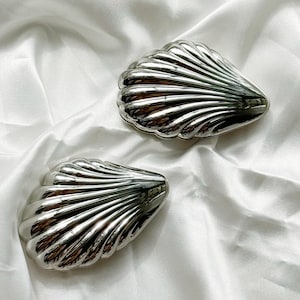 Puede incluir: Dos objetos decorativos plateados con forma de concha brillante. Los objetos tienen una textura acanalada y est&aacute;n colocados sobre un fondo blanco y sedoso. Las conchas reflejan la luz, creando reflejos brillantes.