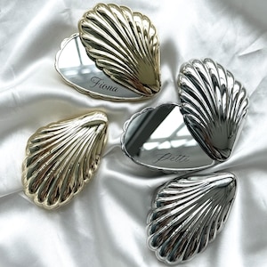 May include: Four shell-shaped compact mirrors in gold and silver finishes. Two mirrors are closed, showing ribbed metallic surfaces. Two mirrors are open, revealing reflective surfaces with the engraved names Fiona and Petti in script font.
