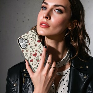 May include: A phone case with a cream background featuring a pattern of red hearts, vines, and flowers. The case is held in front of a person wearing a black leather jacket and a white polka dot shirt.