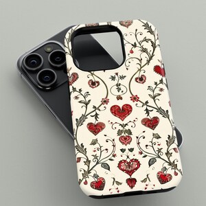 May include: A phone case with a cream background featuring a repeating pattern of red hearts, flowers, and vines. The case has a black border and is shown resting on a black phone with multiple camera lenses.