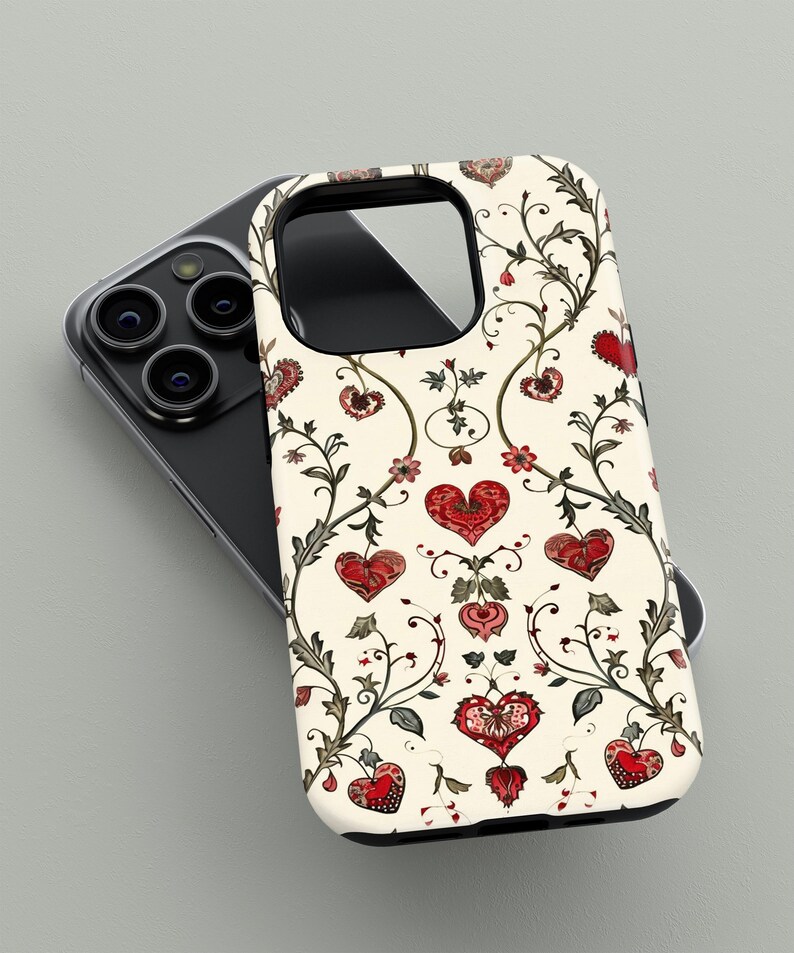 May include: A phone case with a cream background featuring a repeating pattern of red hearts, flowers, and vines. The case has a black border and is shown resting on a black phone with multiple camera lenses.
