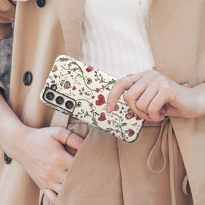 May include: A phone case with a cream background, adorned with a repeating pattern of red hearts, flowers, and vines. The case is held in front of a beige trench coat.
