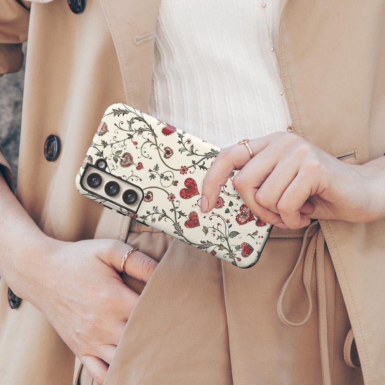 May include: A phone case with a cream background, adorned with a repeating pattern of red hearts, flowers, and vines. The case is held in front of a beige trench coat.