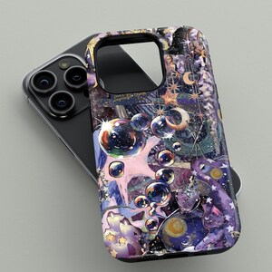 May include: A phone case with a colorful, artistic design featuring bubbles, stars, and celestial elements in shades of purple, pink, and gold. The case is shown on top of a black phone.