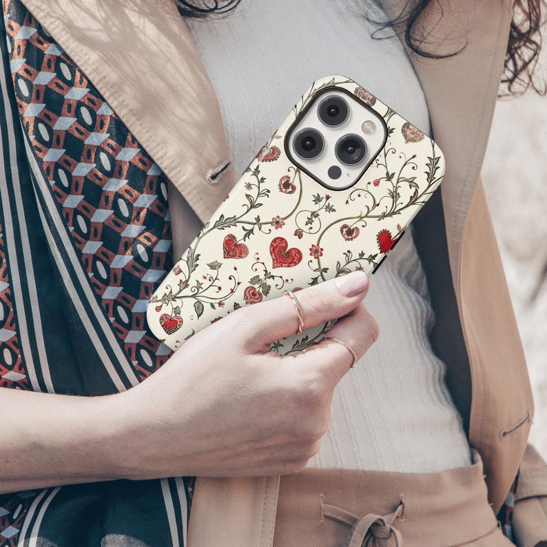 May include: A phone case with a cream background and a repeating pattern of red hearts and green vines. The case has a camera cutout and is held by a person. The case has a decorative floral design.