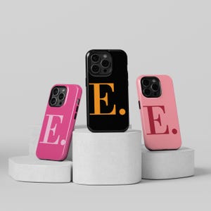 Op de afbeelding: Drie telefoonhoesjes in verschillende roze en zwarte tinten, elk met de letter "E" in een contrasterende kleur. De hoesjes zijn geplaatst op witte sokkels tegen een neutrale achtergrond, wat een minimalistisch ontwerp laat zien.